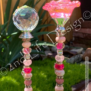 May include: Two pink and white beaded garden stakes with a pink flower and a clear crystal on top. The stakes have pink and white beads, silver spacers, and a pink ribbon charm.