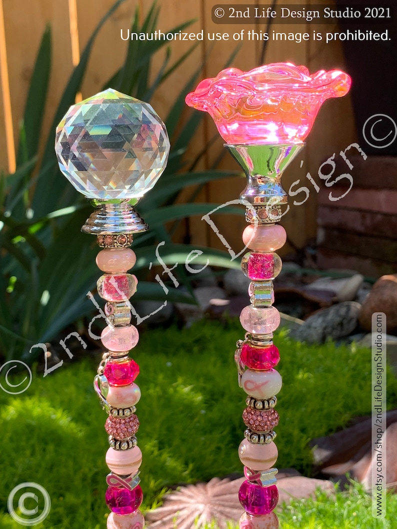 May include: Two pink and white beaded garden stakes with a pink flower and a clear crystal on top. The stakes have pink and white beads, silver spacers, and a pink ribbon charm.
