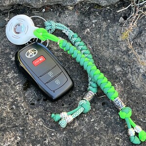 May include: A green and black braided key chain with a silver charm and a black car key with three buttons. The key chain has a white round charm with a design on it.