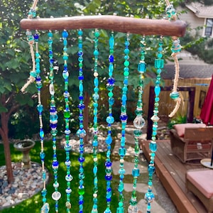 May include: A beaded wind chime with a natural wood branch base. The beads are in shades of blue and green, with some clear glass beads and silver accents. The wind chime is hanging from a brown twine string.
