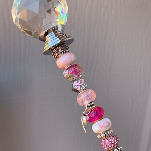 May include: A crystal wand with a silver base and pink and white beads. The beads have pink ribbons and charms, including a pink ribbon charm and a silver angel wing charm.