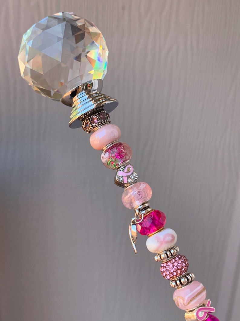 May include: A crystal wand with a silver base and pink and white beads. The beads have pink ribbons and charms, including a pink ribbon charm and a silver angel wing charm.