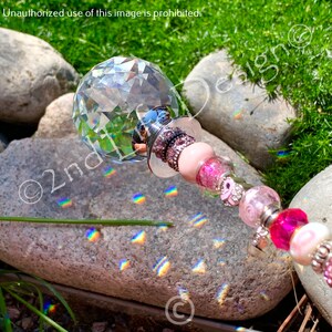 May include: A crystal ball with a pink and silver beaded wand. The wand is decorated with pink and silver beads, and the crystal ball is reflecting rainbows on a gray rock.