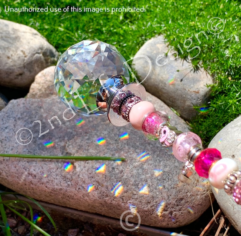 May include: A crystal ball with a pink and silver beaded wand. The wand is decorated with pink and silver beads, and the crystal ball is reflecting rainbows on a gray rock.