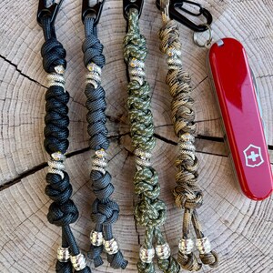 May include: Four keychains with different colored paracord and silver beads. The keychains are attached to black carabiners. A red pocket knife with a white cross is in the background.