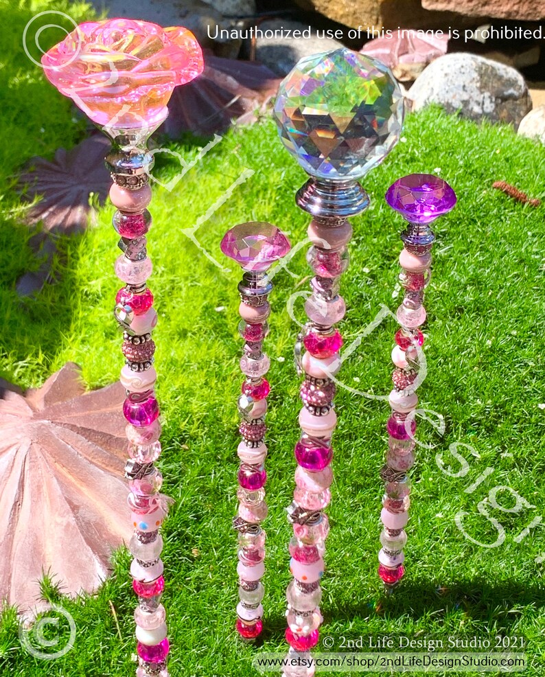 May include: Four decorative garden stakes with pink, white, and clear beads. The top of each stake features a different topper: a pink flower, a clear crystal ball, a pink crystal, and a purple crystal.