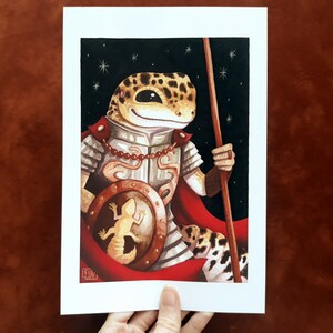 May include: A watercolor painting of a gecko wearing armor and holding a spear. The gecko is wearing a red cape and has a shield with a smaller gecko on it. The background is a dark blue with stars.