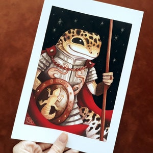 May include: A cartoon illustration of a gecko wearing armor and a red cape, holding a spear and a shield with a smaller gecko on it. The background is a dark blue with white stars.