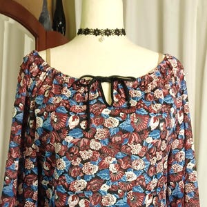 Puede incluir: Una blusa de manga larga con un estampado floral en tonos burdeos, azul y blanco. La blusa tiene un escote redondo con un detalle de lazo negro y bandas negras en los pu&ntilde;os y el dobladillo. La blusa se exhibe en un maniqu&iacute;.