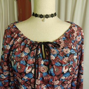 Puede incluir: Una blusa con estampado floral con cuello redondo y mangas largas. La blusa presenta un detalle de lazo negro en el escote y se combina con un collar de gargantilla de encaje negro con un colgante de perla. El estampado floral incluye flores rojas, azules y blancas.