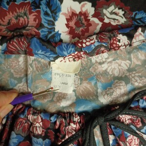 Puede incluir: Primer plano de una prenda con estampado floral con una etiqueta. La tela presenta una mezcla de flores rojas, azules y blancas sobre un fondo oscuro. La etiqueta dice "9615-334", "N", "LARGE" y "5378".
