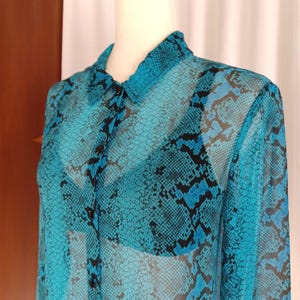 May include: A sheer turquoise button-down shirt with a black snakeskin pattern. The long-sleeved blouse has a collared neckline and black buttons down the front. The fabric appears lightweight and semi-transparent.
