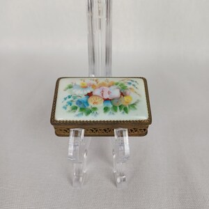 May include: A rectangular, antique trinket box with a floral design. The box features a light green background with a painted bouquet of colorful flowers. The box has a gold-colored metal frame with a decorative, openwork design.