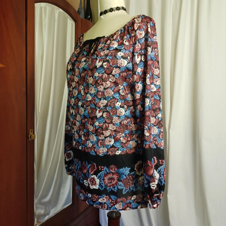 Puede incluir: Una blusa de manga larga con un estampado floral en tonos burdeos, azul y blanco. La blusa tiene una banda negra en la parte inferior y un lazo negro en el escote. La tela parece ligera.