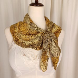 May include: A yellow and brown snakeskin-patterned scarf is draped around a mannequin's neck. The scarf is knotted at the front, showcasing the detailed print. The background is a white curtain.