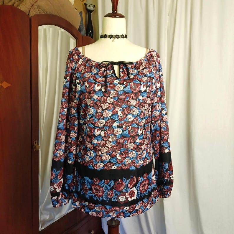 Puede incluir: Una blusa de manga larga con un estampado floral en tonos burdeos, azul y blanco. La blusa tiene un escote redondo con un detalle de lazo negro y bandas negras en los pu&ntilde;os y el dobladillo. La blusa se exhibe en un maniqu&iacute;.