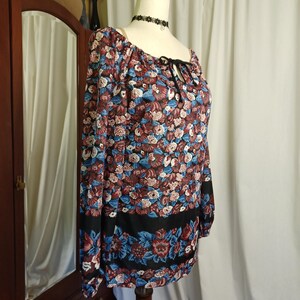 Puede incluir: Una blusa de manga larga con un estampado floral en tonos burdeos, azul y blanco. La blusa tiene un escote cuadrado con un lazo negro y una banda negra en la parte inferior. La blusa se exhibe en un maniqu&iacute;.