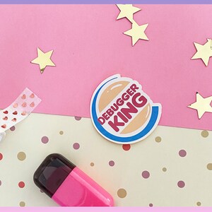 Peut inclure: Un autocollant rose et blanc avec le texte "Debugger King" dans un cercle avec une morsure. L'autocollant est sur un fond rose et beige avec des étoiles dorées et un rouleau de ruban rose et or en forme de cœur.