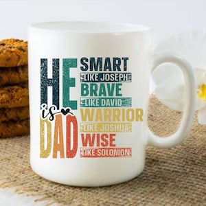 May include: White ceramic mug with the text "He is Dad" in a colorful, vintage-style font. The mug also lists qualities like "Smart," "Brave," "Warrior," and "Wise." A stack of chocolate chip cookies sits to the left of the mug.