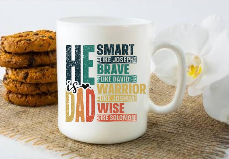 May include: White ceramic mug with the text "He is Dad" in a colorful, vintage-style font. The mug also lists qualities like "Smart," "Brave," "Warrior," and "Wise." A stack of chocolate chip cookies sits to the left of the mug.