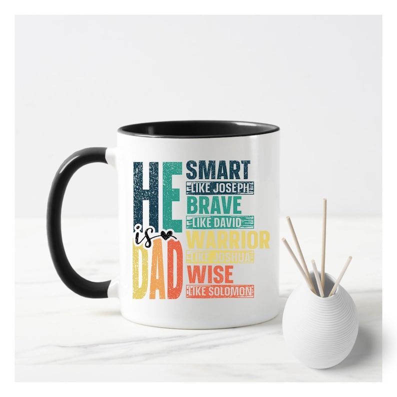 May include: White ceramic mug with a black handle and rim. The mug features the text "HE is DAD" in a colorful, distressed font. Additional text includes "SMART like Joseph", "BRAVE like David", "WARRIOR like Joshua", and "WISE like Solomon".