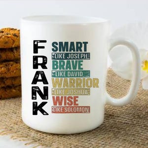 May include: White ceramic mug with the name "FRANK" in large black letters. The mug features the words "SMART," "BRAVE," "WARRIOR," and "WISE" in various colors. A stack of cookies is on the left.