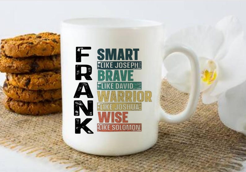May include: White ceramic mug with the name "FRANK" in large black letters. The mug features the words "SMART," "BRAVE," "WARRIOR," and "WISE" in various colors. A stack of cookies is on the left.