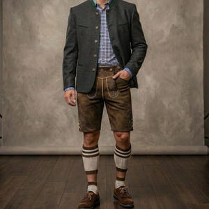 Puede incluir: Traje bávaro tradicional. Chaqueta gris oscuro con ribetes verdes, camisa a cuadros azul y blanco y pantalones cortos de cuero marrón. El conjunto incluye zapatos marrones, calcetines a rayas, sobre un fondo neutro.