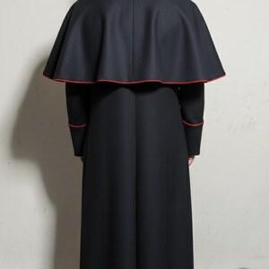 May include: A long, black clerical robe with a matching cape. The cape and cuffs are trimmed with a thin red border. The garment is worn by a person facing away from the camera.