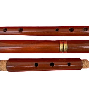 May include: Three-piece wooden flute with a rich, dark brown finish. The flute has brass-colored accents at the ends and around the middle section. The instrument features several finger holes for playing and a cork stopper on one end.