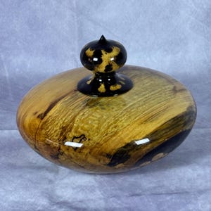 May include: A handcrafted wooden bowl with a lid. The bowl is oval-shaped, featuring a natural wood grain pattern in shades of gold and brown. The lid is topped with a black finial with a gold spotted design. The bowl has a glossy finish.