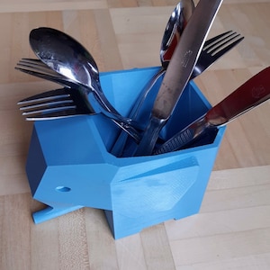 Peut inclure: Un porte-ustensiles bleu en forme d'éléphant rempli d'argenterie. Le support est en plastique et présente un design géométrique. L'argenterie comprend des fourchettes, des cuillères et des couteaux. Le support est posé sur une surface en bois brun clair.