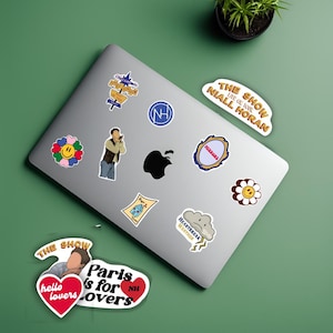 May include: A silver laptop covered in various stickers. The stickers feature a flower with a smiley face, a cloud with "Heartbreak Weather" text, and a sticker that says "The Show Live of You Niall Horan".