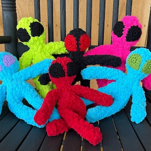 May include: Six crocheted plush alien toys in various colors, including green, pink, blue, red, and black, sitting on a black wooden rocking chair.