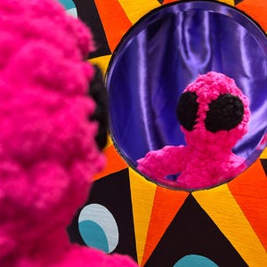 May include: A pink crocheted plush toy with black eyes looks into a round mirror with a purple background. The mirror is set into a colorful, geometric frame with black, yellow, orange, and blue accents.