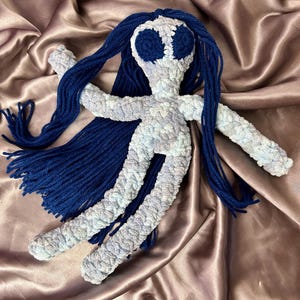 May include: A crocheted doll with long blue hair and a light blue and white body. The doll has large blue eyes and a simple, friendly expression.