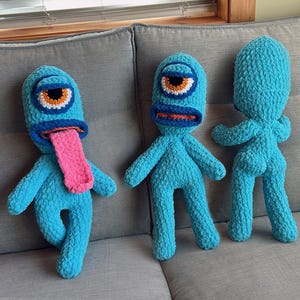 May include: Three handmade blue monster plush toys with one eye, orange and white details, and a pink tongue. The plush toys are on a gray couch. One monster has its tongue out.