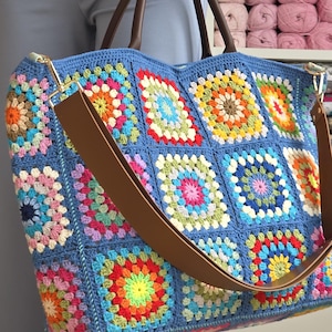 May include: A large, handmade crochet tote bag in shades of blue, with a patchwork design of colorful squares. The bag features brown leather handles and a matching shoulder strap. The bag is filled with a variety of colorful squares.