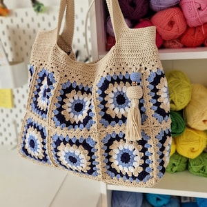 May include: A crocheted tote bag with a blue, white, and beige geometric pattern. The bag has a tassel and is hanging on a hook.