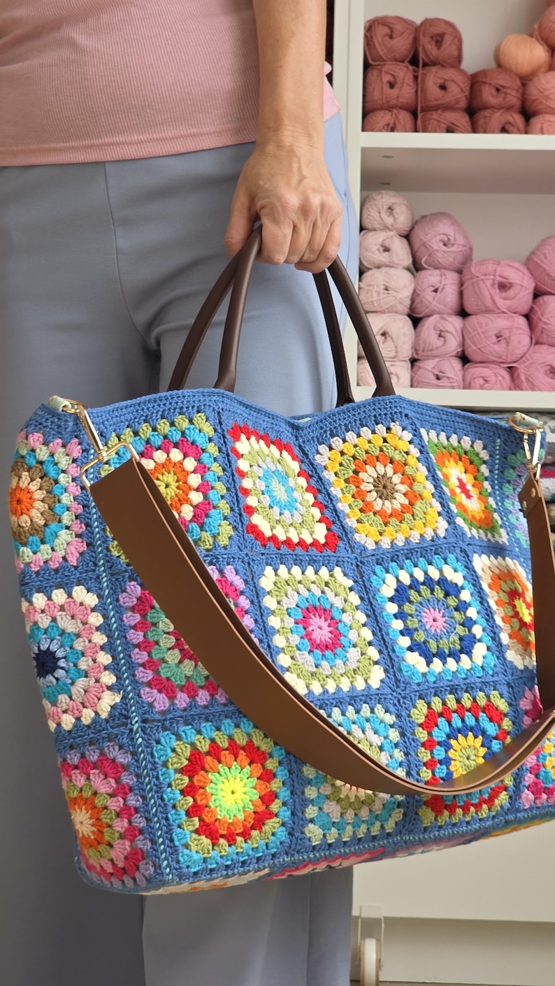 May include: A large, handmade crochet tote bag in shades of blue, with a patchwork design of colorful squares. The bag features brown leather handles and a matching shoulder strap. The bag is filled with a variety of colorful squares.