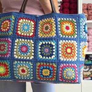 May include: A large, blue crocheted tote bag with a colorful granny square design. The bag features brown leather handles and is held by a person. The squares are in various colors, including red, yellow, green, and pink.