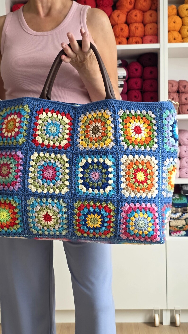 May include: A large, blue crocheted tote bag with a colorful granny square design. The bag features brown leather handles and is held by a person. The squares are in various colors, including red, yellow, green, and pink.