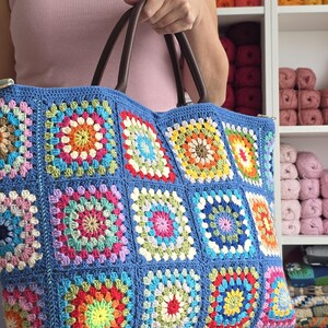 May include: A large, handmade tote bag with a blue crocheted exterior. The bag features colorful square patterns in various shades, including red, yellow, green, and pink. It has brown leather handles and a zipper closure. The bag is held by a person.