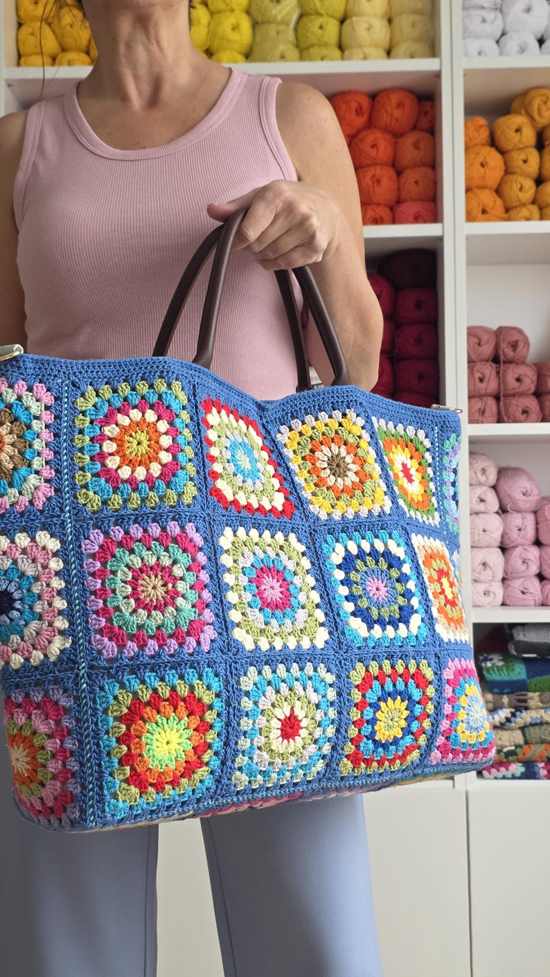 May include: A large, handmade tote bag with a blue crocheted exterior. The bag features colorful square patterns in various shades, including red, yellow, green, and pink. It has brown leather handles and a zipper closure. The bag is held by a person.