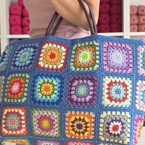 May include: A large, handmade tote bag with a blue background and colorful crochet squares. The squares feature various colors, including orange, green, yellow, and purple. The bag has brown leather handles.