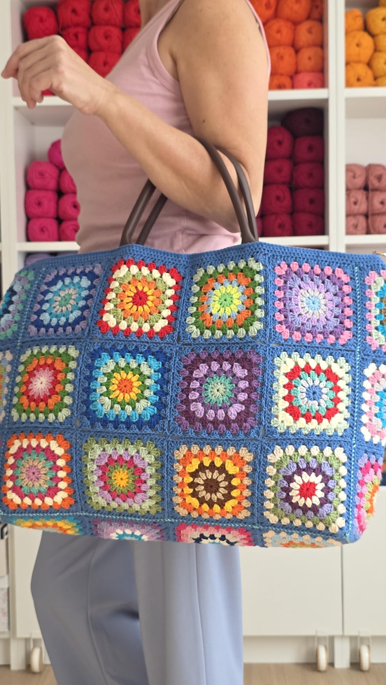 May include: A large, handmade tote bag with a blue background and colorful crochet squares. The squares feature various colors, including orange, green, yellow, and purple. The bag has brown leather handles.