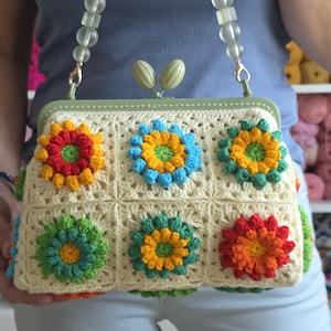 May include: Cream-colored crochet handbag with colorful flower appliqués. The bag features a green frame and a strap made of green beads and clear, round beads. The flowers are in shades of red, orange, yellow, blue, and green.