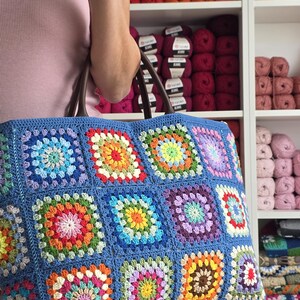 May include: A large, blue crocheted tote bag with a colorful granny square design. The bag features a variety of bright colors, including red, orange, yellow, green, and purple. The bag has brown handles and is held by a person.
