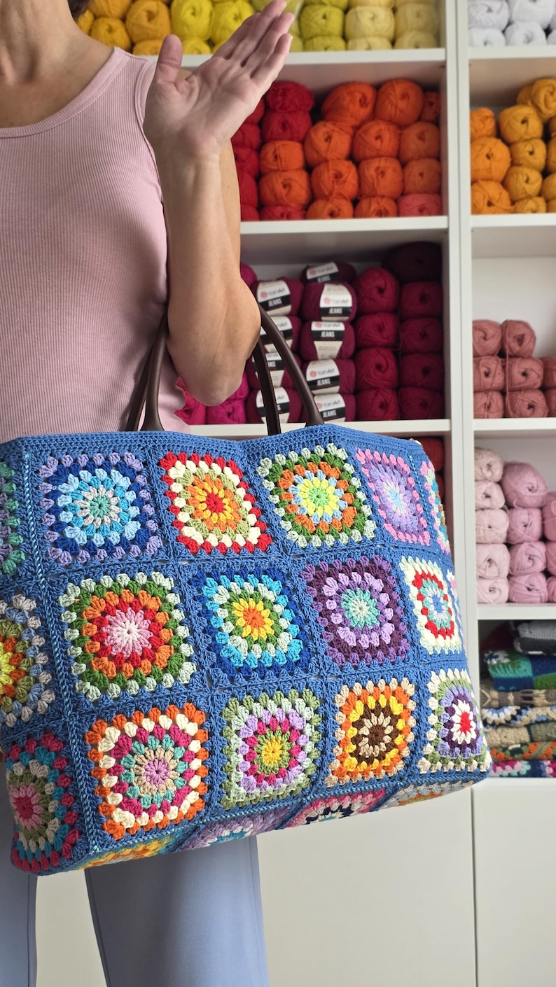 May include: A large, blue crocheted tote bag with a colorful granny square design. The bag features a variety of bright colors, including red, orange, yellow, green, and purple. The bag has brown handles and is held by a person.