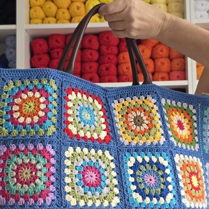 May include: A blue crocheted tote bag with a colorful granny square design. The bag features brown leather handles and is filled with a variety of yarn colors in the background.